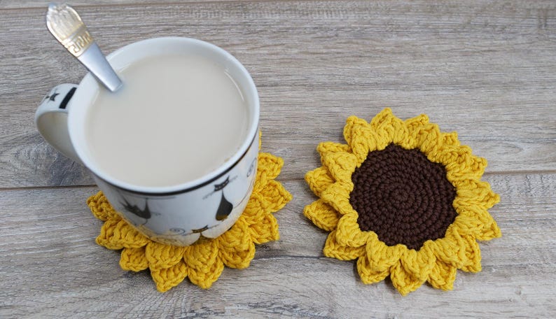 May include: A cup of tea with a spoon rests on a yellow crochet sunflower coaster. A second sunflower coaster is next to the cup. The coasters have a brown center and yellow petals. The cup has a black cat design.