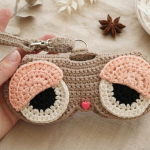 K&ouml;nnte beinhalten: Ein beiges Brillenetui aus H&auml;kelarbeit mit zwei gro&szlig;en Augen, bestehend aus schwarzen Pupillen, wei&szlig;en Iris und rosafarbenen Lidern. Das Etui besitzt eine passende beige H&auml;kelschlaufe mit silbernem Metallverschluss und eine kleine rosa Herzperle.