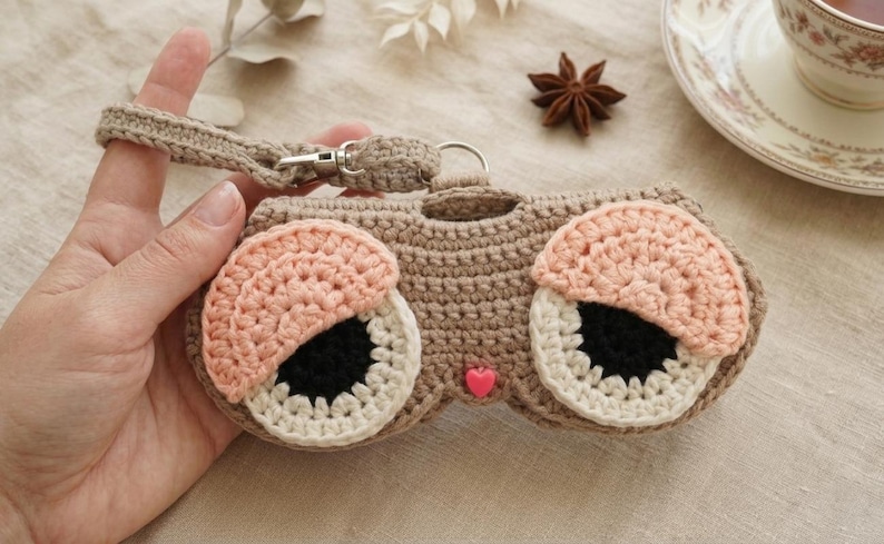 K&ouml;nnte beinhalten: Ein beiges Brillenetui aus H&auml;kelarbeit mit zwei gro&szlig;en Augen, bestehend aus schwarzen Pupillen, wei&szlig;en Iris und rosafarbenen Lidern. Das Etui besitzt eine passende beige H&auml;kelschlaufe mit silbernem Metallverschluss und eine kleine rosa Herzperle.