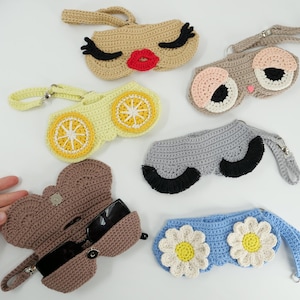 K&ouml;nnte beinhalten: Sechs geh&auml;kelte Brillenetuis mit Handschlaufen. Die Designs umfassen ein Gesicht mit roten Lippen und schwarzen Wimpern, ein Gesicht mit gro&szlig;en Augen, ein gelbes Zitronenscheibenmuster, ein graues Etui mit schwarzen Wimpern, ein braunes Etui und blaue G&auml;nsebl&uuml;mchen.