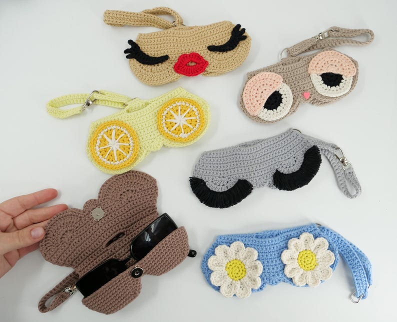 K&ouml;nnte beinhalten: Sechs geh&auml;kelte Brillenetuis mit Handschlaufen. Die Designs umfassen ein Gesicht mit roten Lippen und schwarzen Wimpern, ein Gesicht mit gro&szlig;en Augen, ein gelbes Zitronenscheibenmuster, ein graues Etui mit schwarzen Wimpern, ein braunes Etui und blaue G&auml;nsebl&uuml;mchen.