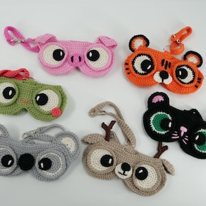May include: A collection of six crocheted eyeglass cases in the shape of animals. The cases include a pink pig, orange tiger, green frog, gray koala, brown deer, and black cat. Each case has large, round eyes and a strap.