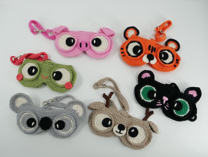 May include: A collection of six crocheted eyeglass cases in the shape of animals. The cases include a pink pig, orange tiger, green frog, gray koala, brown deer, and black cat. Each case has large, round eyes and a strap.
