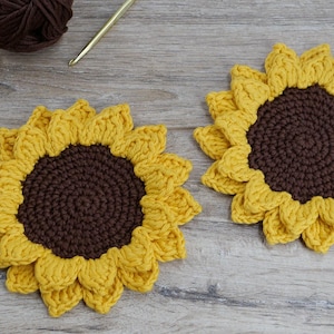 May include: Two handmade crochet sunflower coasters with yellow petals and brown centers. The coasters are placed on a wooden surface. Yellow and brown yarn and a crochet hook are in the background.