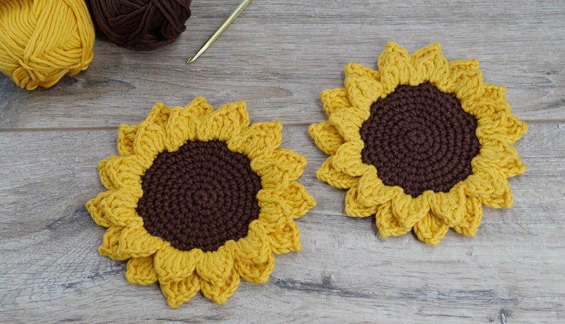May include: Two handmade crochet sunflower coasters with yellow petals and brown centers. The coasters are placed on a wooden surface. Yellow and brown yarn and a crochet hook are in the background.