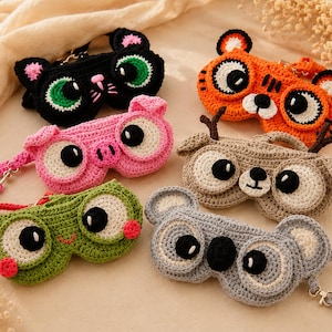 May include: A collection of handmade crochet eyeglass cases in various animal designs. The cases include a black cat, orange tiger, pink pig, tan deer, green frog, and gray koala. Each case features large, expressive eyes and a strap.