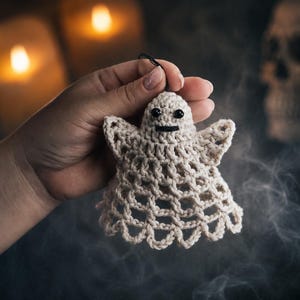 Op de afbeelding: Een klein, gebroken wit gehaakt spookornament met een delicaat, opengewerkt rasterpatroon over het hele lichaam. Het spookje heeft twee kleine, ronde zwarte kraaloogjes en een eenvoudig, recht zwart geborduurd lijntje als mond.