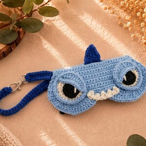 May include: A light blue crocheted eyeglass case shaped like a cartoon monster with large eyes, a fin, and teeth. It has a matching blue strap with a silver clasp for easy carrying. The case is designed to protect eyeglasses.