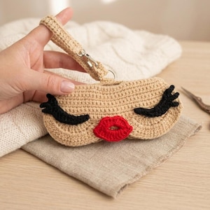 Peut inclure: Une pochette en crochet beige arborant un visage avec des cils en laine noire et des lèvres en laine rouge vif. La pochette comprend une dragonne en crochet beige assortie fixée par un fermoir pivotant en métal argenté et un anneau.