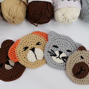 Op de afbeelding: Vier gehaakte dierenonderzetters, waaronder een bruine beer, een beige beer met oranje oren, een grijze kat en een beige kat. De onderzetters zijn in een halve cirkel op een wit oppervlak gerangschikt.
