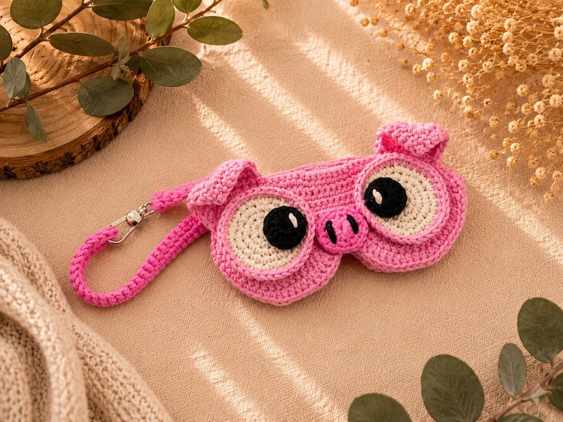 May include: A pink crocheted sleep mask shaped like a pig, featuring large white and black eyes, a pink snout, and small ears. The mask has a matching pink strap with a silver clasp. The item is handmade and designed for sleep.