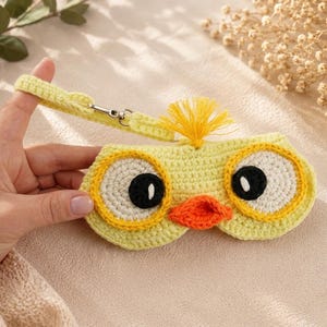 May include: A handmade yellow crochet glasses case shaped like a chick with large eyes, an orange beak, and a yellow tassel. The case has a strap with a metal clasp. The case is held by a hand.