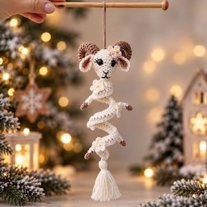 May include: A cream-colored crocheted ram ornament with brown curved horns, black eyes, and pink-centered white flowers. The body is a long, spiraling, cream-colored crocheted tail adorned with small white flowers, ending in a cream-colored yarn tassel.