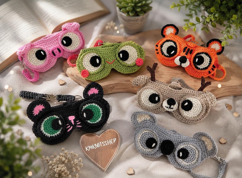 May include: A collection of handmade crochet sleep masks in various animal designs, including a pink pig, green frog, orange tiger, black cat, tan deer, and gray koala. Each mask features large, expressive eyes and is crafted with colorful yarn.
