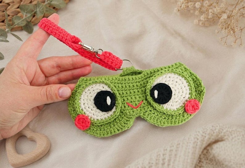 May include: A green crocheted sleep mask shaped like a frog with large white and black eyes, a small orange smile, and pink cheeks. It has a red strap with a silver clasp.