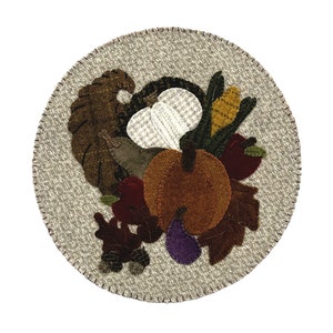 May include: A round, beige placemat with a stitched border, featuring a felt applique of a cornucopia. The cornucopia includes a brown pumpkin, red apples, and various leaves, corn, and other harvest elements.