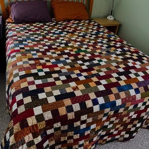 May include: A handmade quilt with a patchwork design in various colors, including red, brown, blue, and cream. The quilt covers a bed with a wooden headboard and two pillows. The bed is in a room with a green wall.