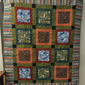 May include: A handmade quilt with a patchwork design. The quilt features nine square blocks in shades of green, orange, and brown. Some blocks have sports-themed patterns, while others have abstract designs. The border has a striped pattern with various colors.