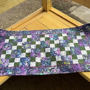 May include: A rectangular quilted table runner with a checkerboard pattern. The squares alternate between white, green, and a floral print in shades of purple and blue. The border features a floral pattern and a thin purple trim.