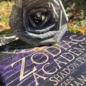 May include: A book titled "Zodiac Academy: The Shadow  Protocol" by Caroline  Cckham. The book cover features a purple brick wall with gold lettering. A black paper rose with a silver star pattern is resting on the book.