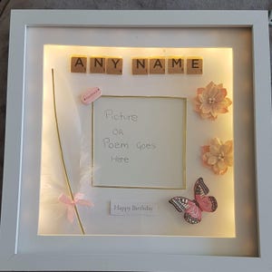 May include: A white picture frame with a light inside. The frame has a white mat and a gold border. The frame is decorated with a feather, a butterfly, and two flowers. The frame also has a small card that says "Happy Birthday". The frame has a space for a picture or poem. The top of the frame has the letters "ANY NAME" spelled out with Scrabble tiles.