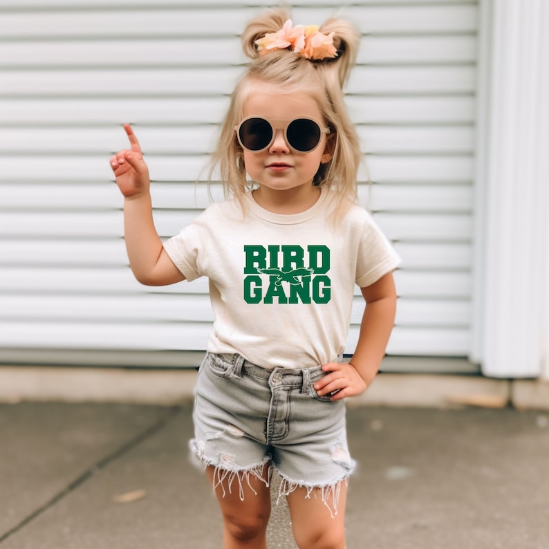 May include: A young girl wearing a cream colored t-shirt with the words "Bird Gang" in green letters. The girl is wearing denim shorts and round sunglasses.