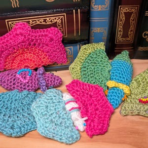 May include: A collection of small, handmade crochet appliques in various shapes and colours, including bright pink, teal, light blue, lime green, and purple. Some pieces feature small, colourful beads sewn onto the textured yarn surfaces.