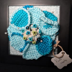May include: A framed, handmade crochet flower in shades of blue and turquoise. The flower is embellished with small beads and pearls. The frame is black, and a small tag with the words "Viola azules" is attached.