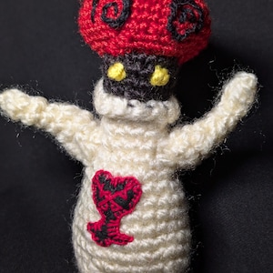 May include: A crocheted plush toy shaped like a mushroom with a red cap and white body. The mushroom has black eyes and a yellow heart on its chest.