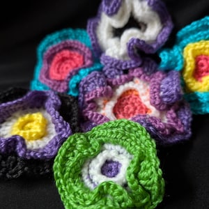 May include: A selection of vibrant, handmade crochet flowers in different sizes and patterns. The flowers display a variety of colours, such as purple, green, yellow, pink, and blue, with white centres. The crochet work is detailed and textured.