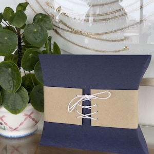 Peut inclure: Un oreiller bleu avec une bande de papier brun attachée avec une ficelle blanche. La bande a un motif de couture décoratif.