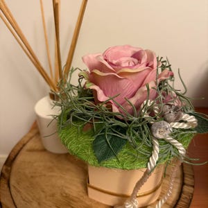 Pode incluir: Uma rosa rosa com folhas verdes e barbante branco está em uma pequena caixa em forma de coração com uma tampa verde e branca. A caixa está sobre uma superfície de madeira.