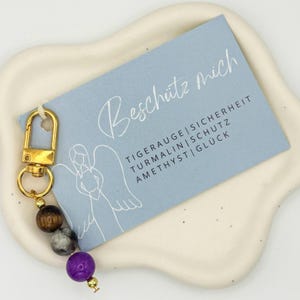 Könnte beinhalten: Ein goldfarbener Schlüsselanhänger mit Karabinerhaken, verziert mit einer Kette aus braunen, grauen und violetten Perlen. Ein hellblaues Etikett mit der Aufschrift "Beschütz mich" und anderem Text ist angebracht.