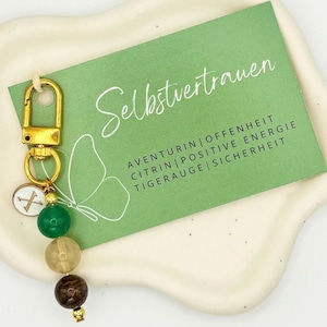 Könnte beinhalten: Ein goldfarbener Schlüsselanhänger mit einem Anhänger und grünen, beigen und braunen Perlen. Eine grüne Karte mit dem Wort "Selbstvertrauen" und anderem Text befindet sich im Hintergrund. Der Schlüsselanhänger hat einen goldenen Verschluss und einen kleinen Anhänger mit einem weißen X.
