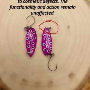 May include: Two purple fishing lures with white spots and silver hooks. The lures are teardrop-shaped and have a shiny finish. Text on the image reads: "Please Note: This item is a clearance item due to cosmetic defects. The functionality and action remain unaffected."