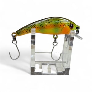 May include: A fishing lure with a yellow and green body, orange accents, and black dots. The lure has two silver hooks and a clear plastic lip. It is displayed on a clear plastic stand against a white background.