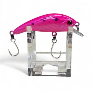 May include: A bright pink fishing lure with dark purple spots and a small silver lip. The lure has two silver hooks and is displayed on a clear acrylic stand. The fishing lure is designed to attract fish.