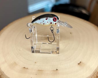 May include: A white and black fishing lure with red spots. The lure is on a clear plastic display stand.