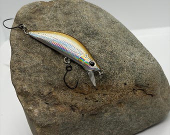 May include: A fishing lure with a metallic gold and silver holographic finish, featuring a black eye, a clear plastic diving lip, and two black metal hooks attached to the body with small silver rings.