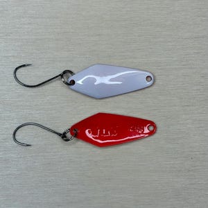 May include: Two fishing lures. One is white, the other is red. Both have a diamond shape with a hook attached. The red lure has the text "4.6g" printed on it. The lures are designed for catching fish.