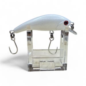 May include: A white fishing lure with a textured body and red eyes is displayed on a clear acrylic stand. The lure has two silver hooks and a small, clear plastic fin. The lure is designed for catching fish.
