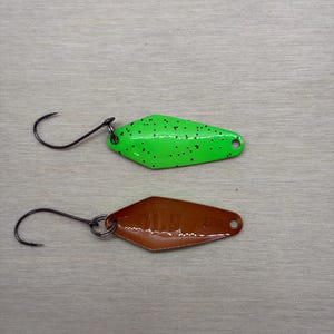 May include: Two fishing lures. The top lure is bright green with black speckles. The bottom lure is brown with a diamond shape and the number "2.09" printed on it. Both lures have a black hook attached.