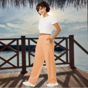 May include: Peach-colored, wide-leg pants with a small pocket detail and the word "XOXO" embroidered on the front. The pants are paired with a white crop top and white sneakers. The outfit is casual and suitable for warm weather.