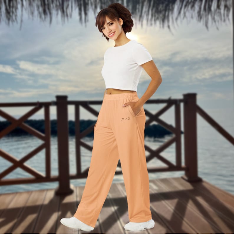 May include: Peach-colored, wide-leg pants with a small pocket detail and the word "XOXO" embroidered on the front. The pants are paired with a white crop top and white sneakers. The outfit is casual and suitable for warm weather.