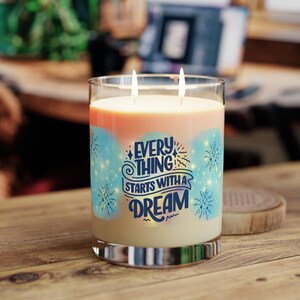 Könnte beinhalten: Eine große Glaskerze mit zwei Dochten, mit einem hellorangen und blauen Design. Die Kerze trägt den Text "EVERYTHING STARTS WITH A DREAM" in blauer Schrift. Die Kerze steht auf einer Holzoberfläche.