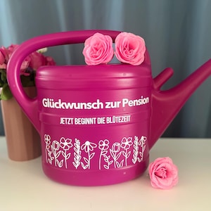 Puede incluir: Una regadera magenta con el texto "Glückwunsch zur Pension" y "JETZT BEGINNT DIE BLÜTEZEIT" en blanco. La regadera tiene un diseño floral y está decorada con rosas rosas. Un jarrón de rosas rosas está en el fondo.