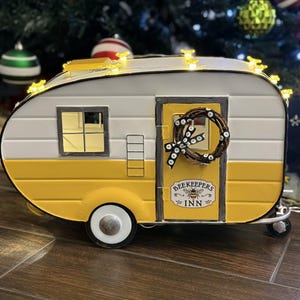 May include: A yellow and white metal camper with a wreath on the door and the text "Beekeepers Inn". The camper has a window and a door. There are lights on the roof of the camper.