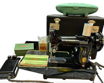 Puede incluir: Máquina de coser Singer negra vintage con pedal, accesorios y maletín de transporte. La máquina tiene detalles dorados y una bobina de hilo blanca. Una funda verde se encuentra encima del maletín negro. El logotipo de Singer es visible.