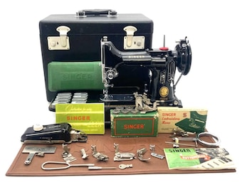 May include: A vintage black Singer sewing machine with a carrying case, accessories, and instruction manuals. The machine has a gold emblem and is surrounded by green and brown boxes and tools. The sewing machine is on a brown mat.