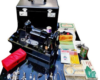 Puede incluir: Una máquina de coser Singer negra vintage con un maletín negro, accesorios y una funda roja. La máquina tiene un carrete de hilo morado y está rodeada de varias herramientas y cajas.